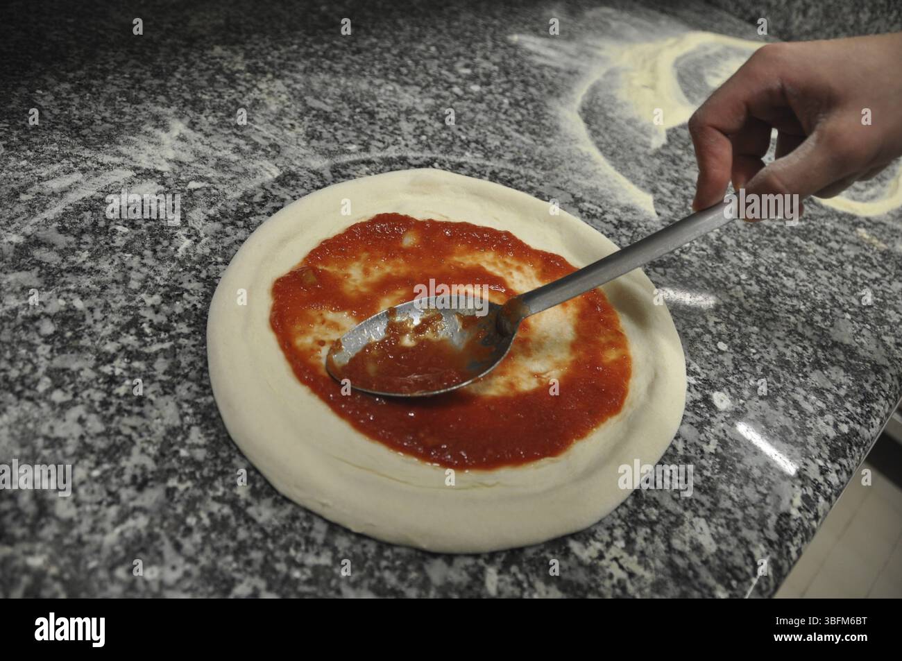 La salsa di pomodoro viene spalmata su una base di pizza con un cucchiaio su una superficie di granito, cuochi e chef preparano la cena con la tipica dieta Mediterranea a l Foto Stock