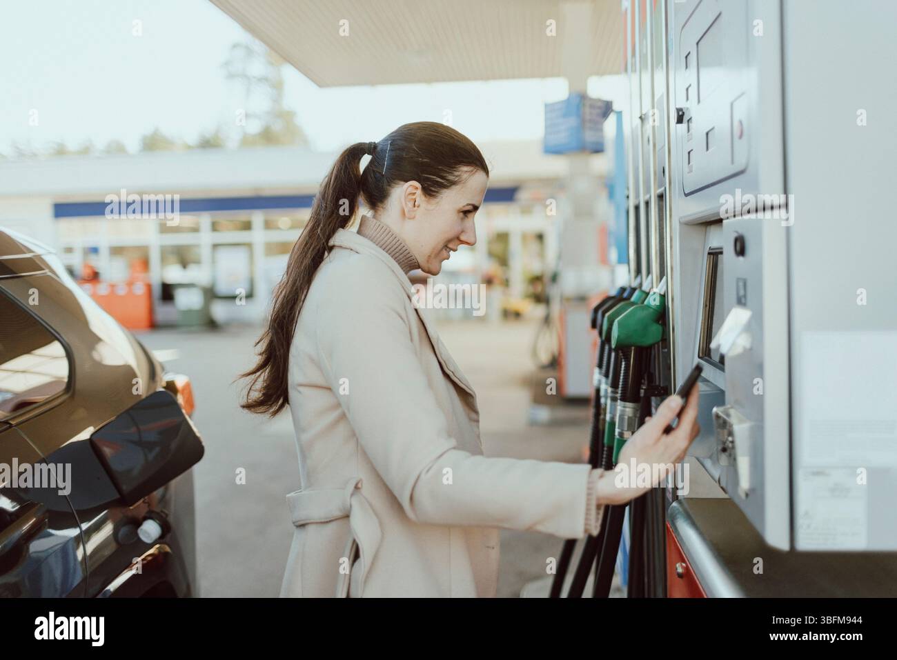 Vista laterale della donna con coda di cavallo che effettua il pagamento online dopo aver rifornito l'auto al distributore di benzina Foto Stock