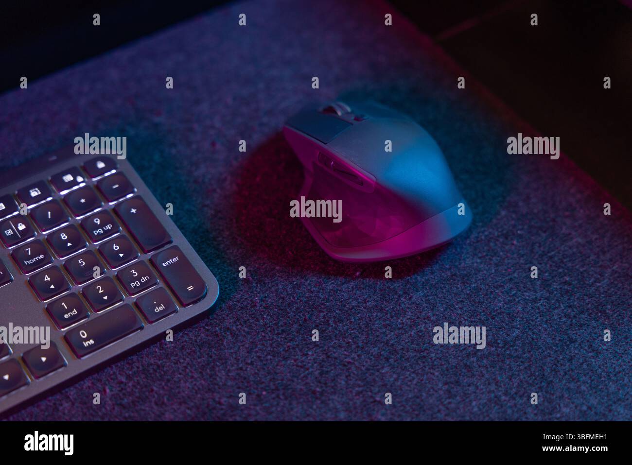 Primo piano dettagliato di un mouse ergonomico per computer e di una parte di una tastiera retroilluminata, immersa in una vivace luce ambiente rosa e blu su un des scuro Foto Stock