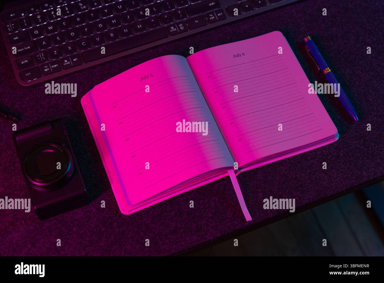 Un pianificatore giornaliero aperto che mostra le date di luglio, accompagnato da una penna, una piccola fotocamera o dispositivo e una tastiera, il tutto sotto una vivace luce di colore rosa. Perf Foto Stock