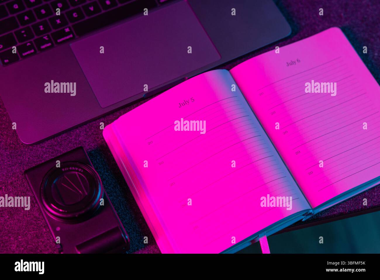 Un piano di lavoro dall'alto con un pianificatore aperto di luglio, un laptop e una piccola fotocamera o dispositivo, il tutto immerso in una calda luce rosa. ID Foto Stock