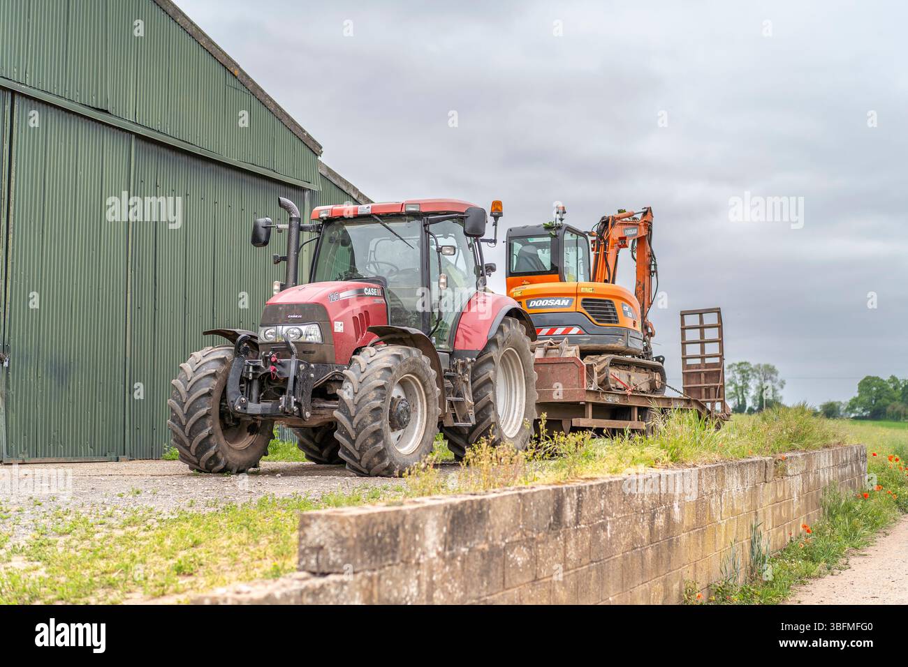 Vista anteriore del trattore agricolo con rimorchio e scavatore sul lato posteriore del rimorchio. Il trattore è parcheggiato davanti a un grande corr verde Foto Stock