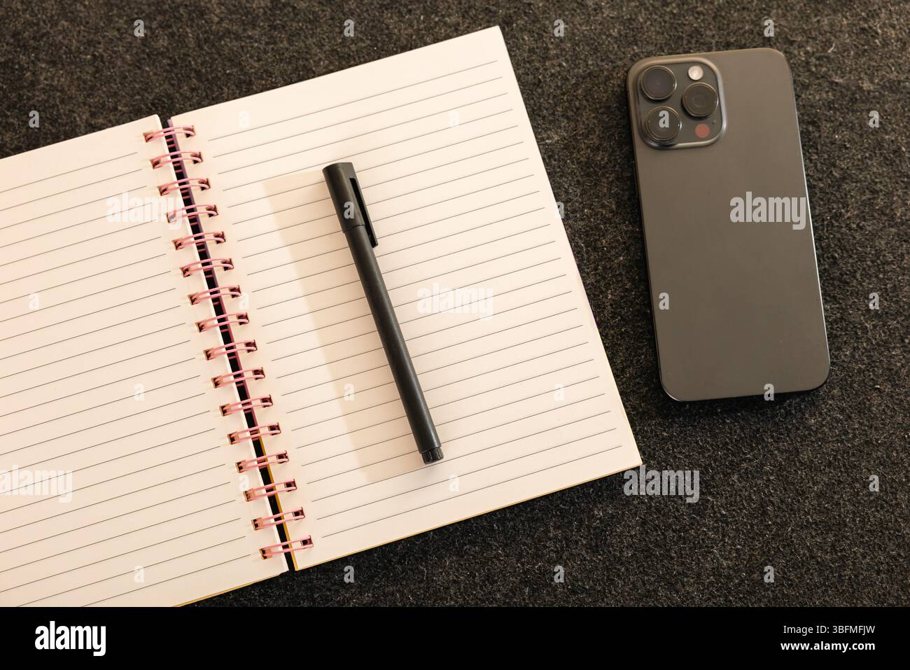 Composizione piatta e minimalista con un notebook a spirale aperto, una penna nera e uno smartphone moderno su un tappetino da scrivania grigio scuro. Ideale Foto Stock