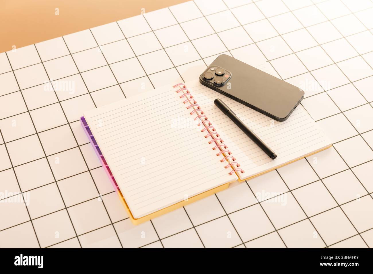 Disposizione piatta top-down di un quaderno a spirale aperto con carta foderata, una penna nera e uno smartphone, disposti su una superficie bianca con griglia nera. T Foto Stock