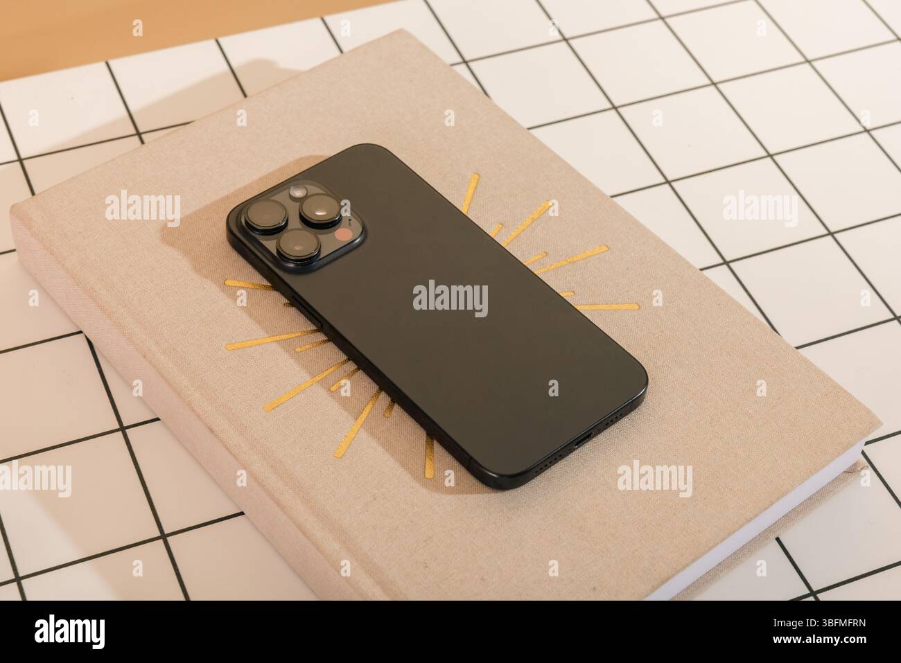 Primo piano di uno smartphone scuro contemporaneo appoggiato su un libro chiuso o un diario con un design dorato Sunburst, su un bianco e un blu scuro Foto Stock