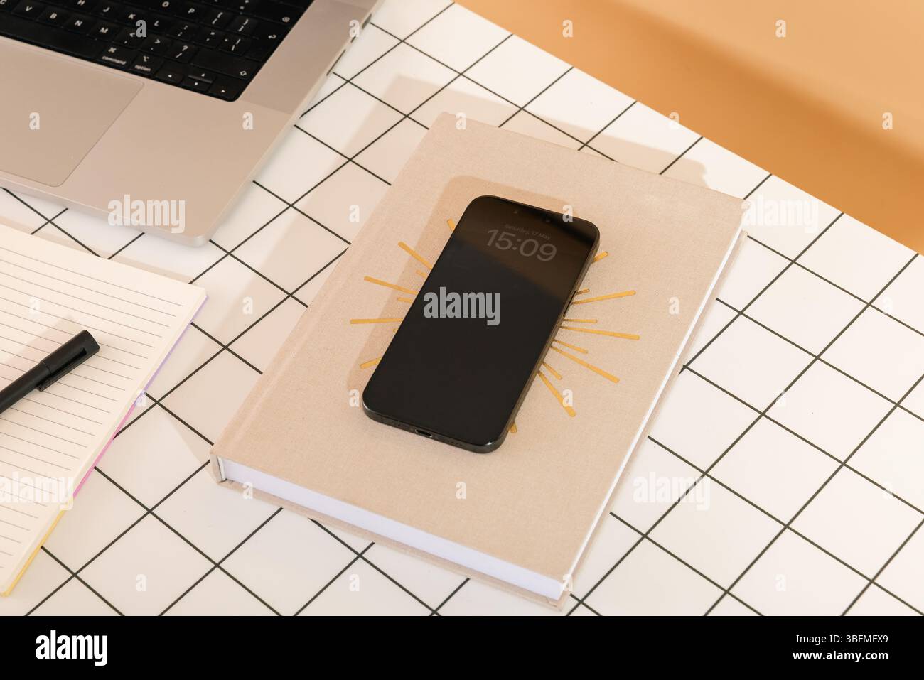 Vista dall'alto di una scrivania organizzata con uno smartphone che mostra l'ora su un libro dal design Sunburst, accanto a un laptop e a un notebook aperto con un p Foto Stock