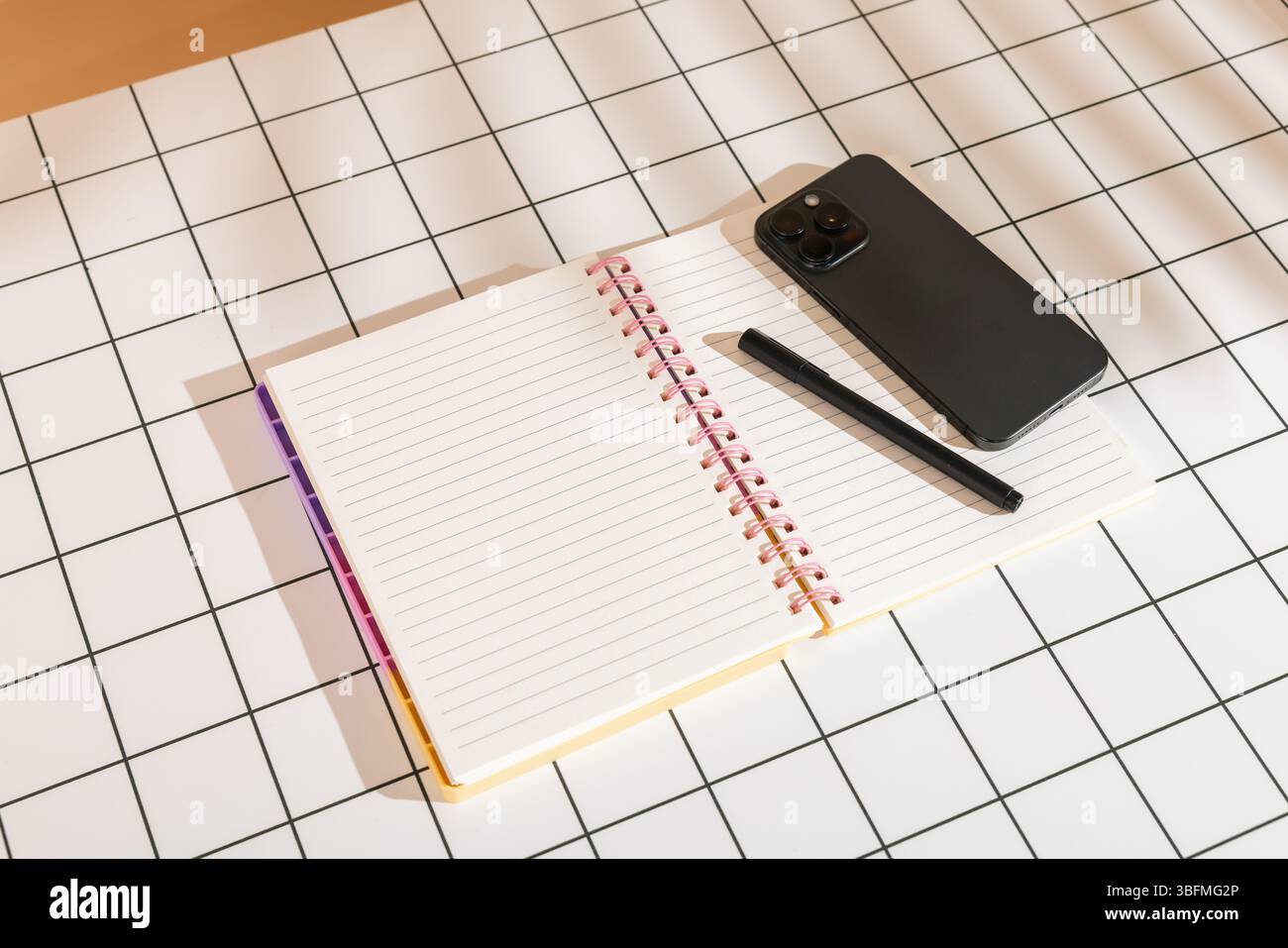 Vista dall'alto ravvicinata di un'area di lavoro organizzata con un notebook aperto, una penna nera e uno smartphone su una griglia bianca e nera Foto Stock