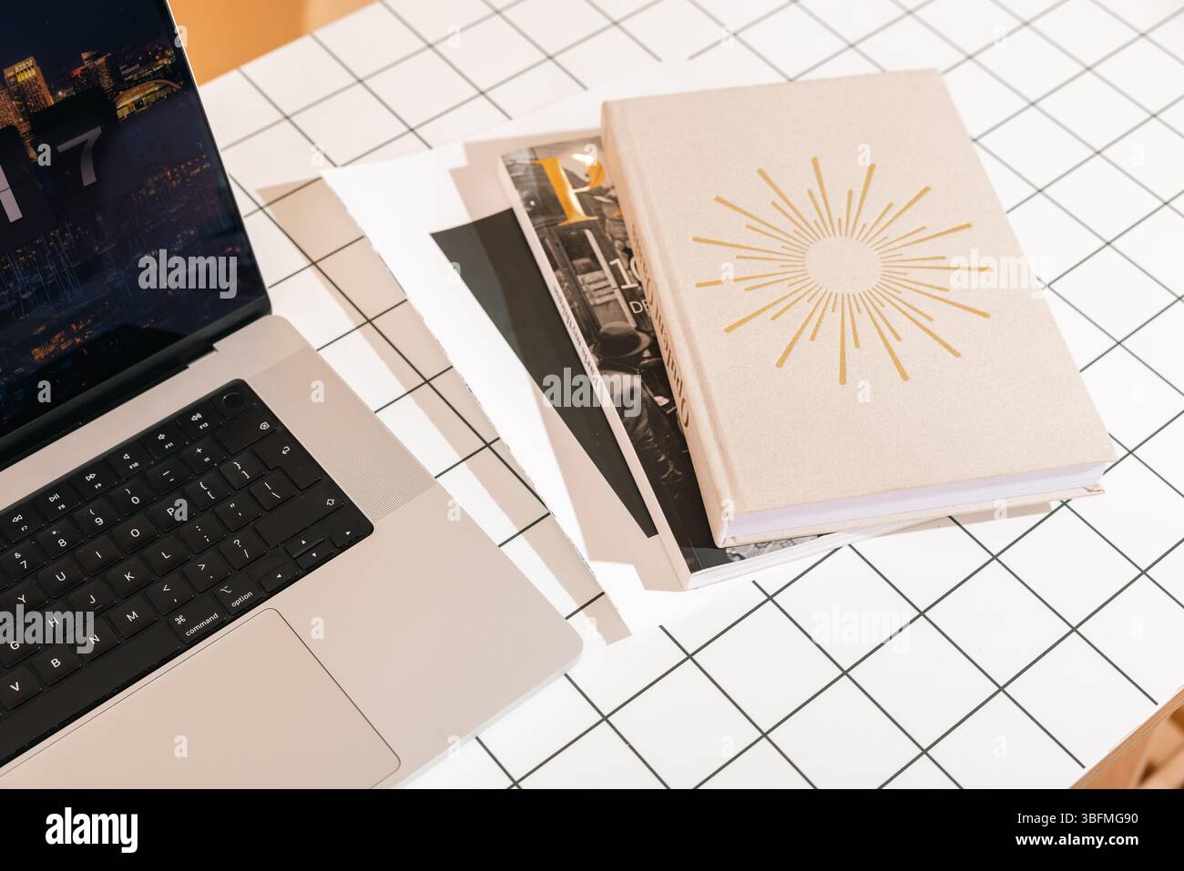 Una vista dall'alto verso il basso di una scrivania elegante e organizzata con un laptop, una pila di riviste o libri (uno con un design Sunburst), su una griglia moderna Foto Stock