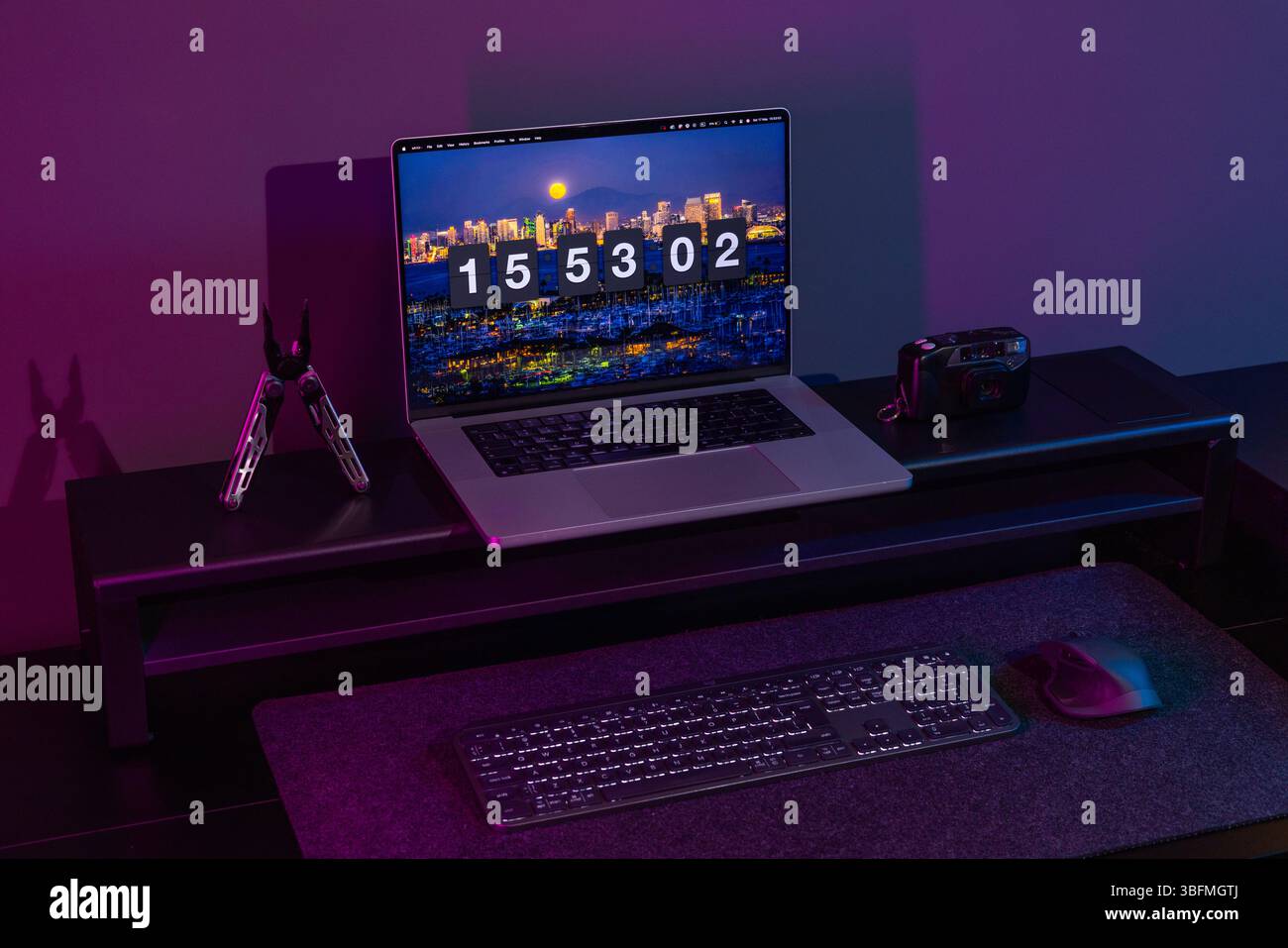 Un'angolazione alternativa di uno spazio di lavoro tecnologico e creativo contemporaneo, che presenta un laptop con screensaver cittadino, una scrivania con multitool e fotocamera Foto Stock