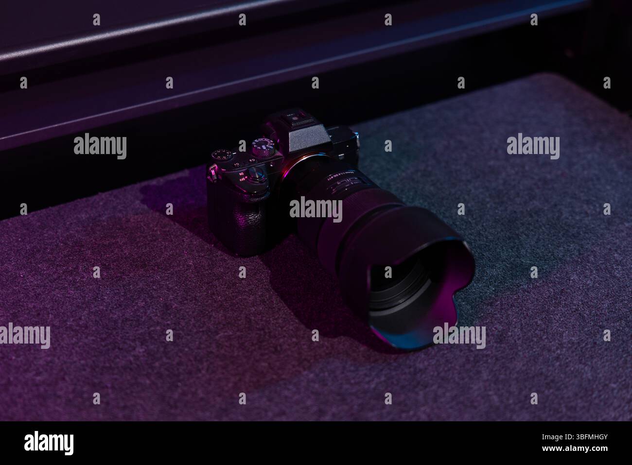 Una moderna fotocamera digitale mirrorless nera con un grande obiettivo collegato, su uno sfondo scuro con sottili riflessi di luce al neon rosa e blu. Foto Stock