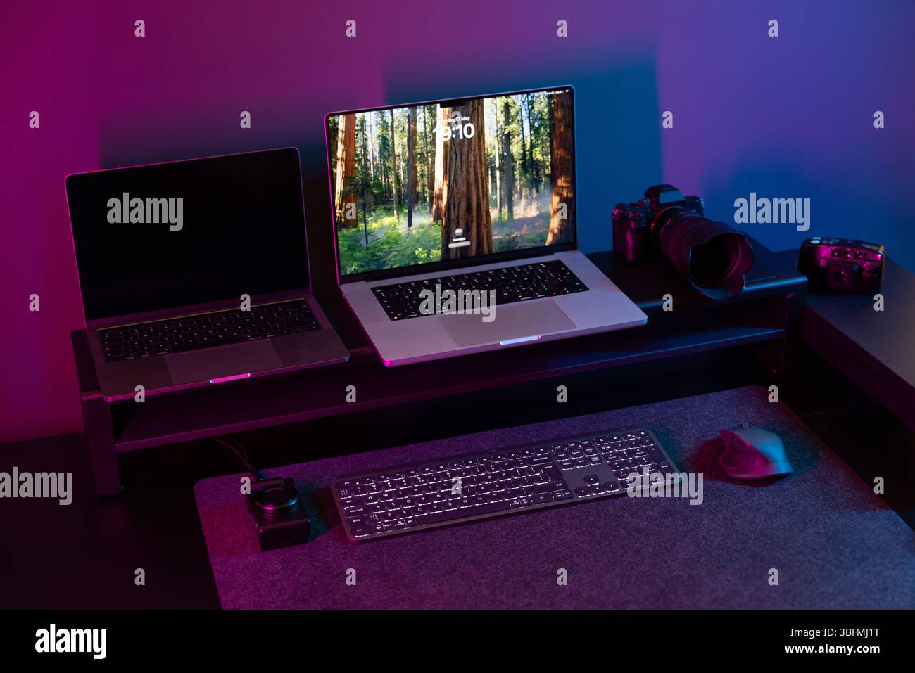 Una moderna configurazione da scrivania con due laptop e una fotocamera su uno scaffale, con un laptop che visualizza uno screensaver forestale. Una tastiera e un mouse sono belli Foto Stock