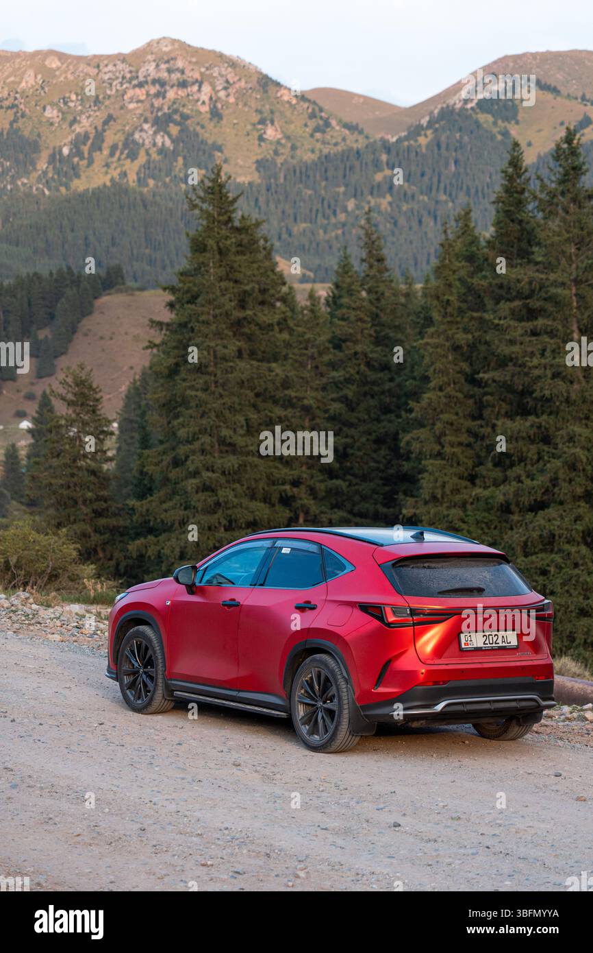 Bishkek, Kirghizistan - 12 ottobre 2024: Vista dall'angolo posteriore di un SUV Lexus NX350 rosso su una strada sterrata, con alberi sempreverdi e montagne sullo sfondo Foto Stock