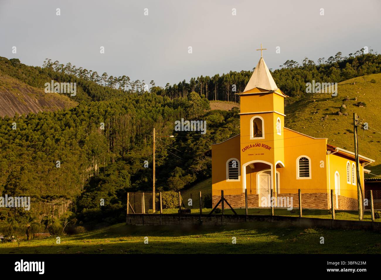 Affascinante cappella di campagna gialla di São Roque illuminata dalla calda luce pomeridiana, circondata da lussureggianti colline verdi e foreste nella campagna brasiliana. Foto Stock