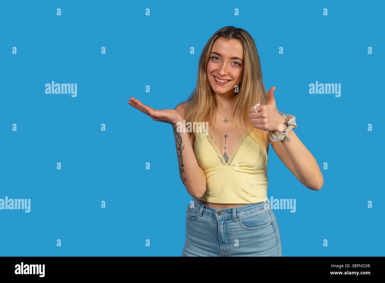 Una donna allegra tatuata che fa un gesto con il pollice e mostra uno spazio vuoto per le copie sul palmo aperto, consigliando qualcosa sullo sfondo blu dello studio Foto Stock