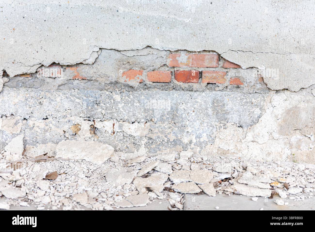 Primo piano di una parete in cemento deteriorata con intonaco sbriciolato che rivela mattoni rossi e detriti sparsi sul terreno. Foto Stock