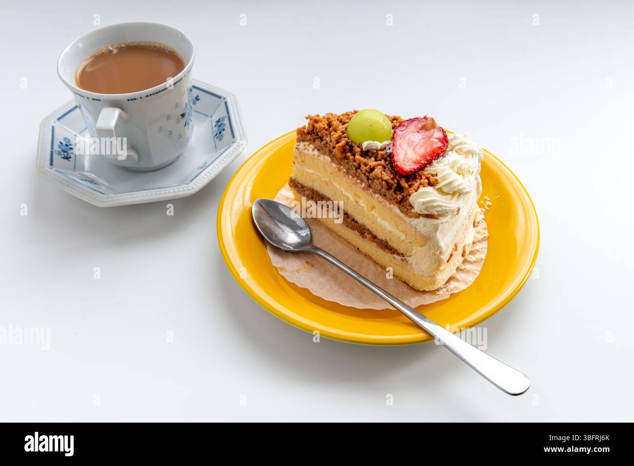 Una fetta di torta alla panna con guarnizioni di frutta servita con una tazza di tè al latte isolato su bianco. Foto Stock