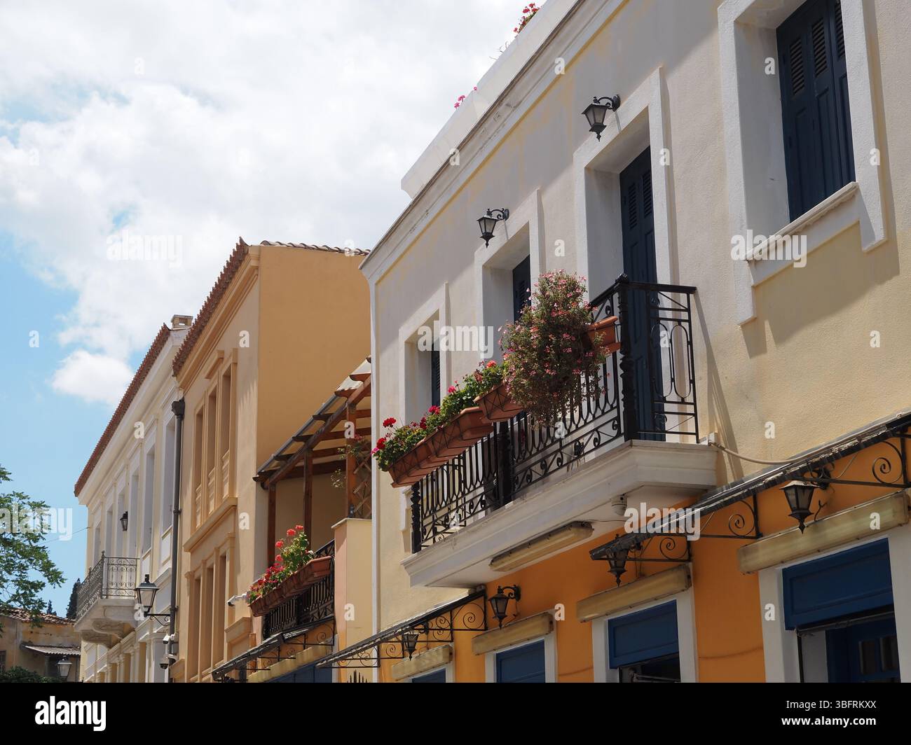 Parti superiori di case mediterranee gialle e arancioni con balconi scuri e fiori rosa sotto un cielo caldo e nuvoloso. Ambiente urbano costiero accogliente. Foto Stock