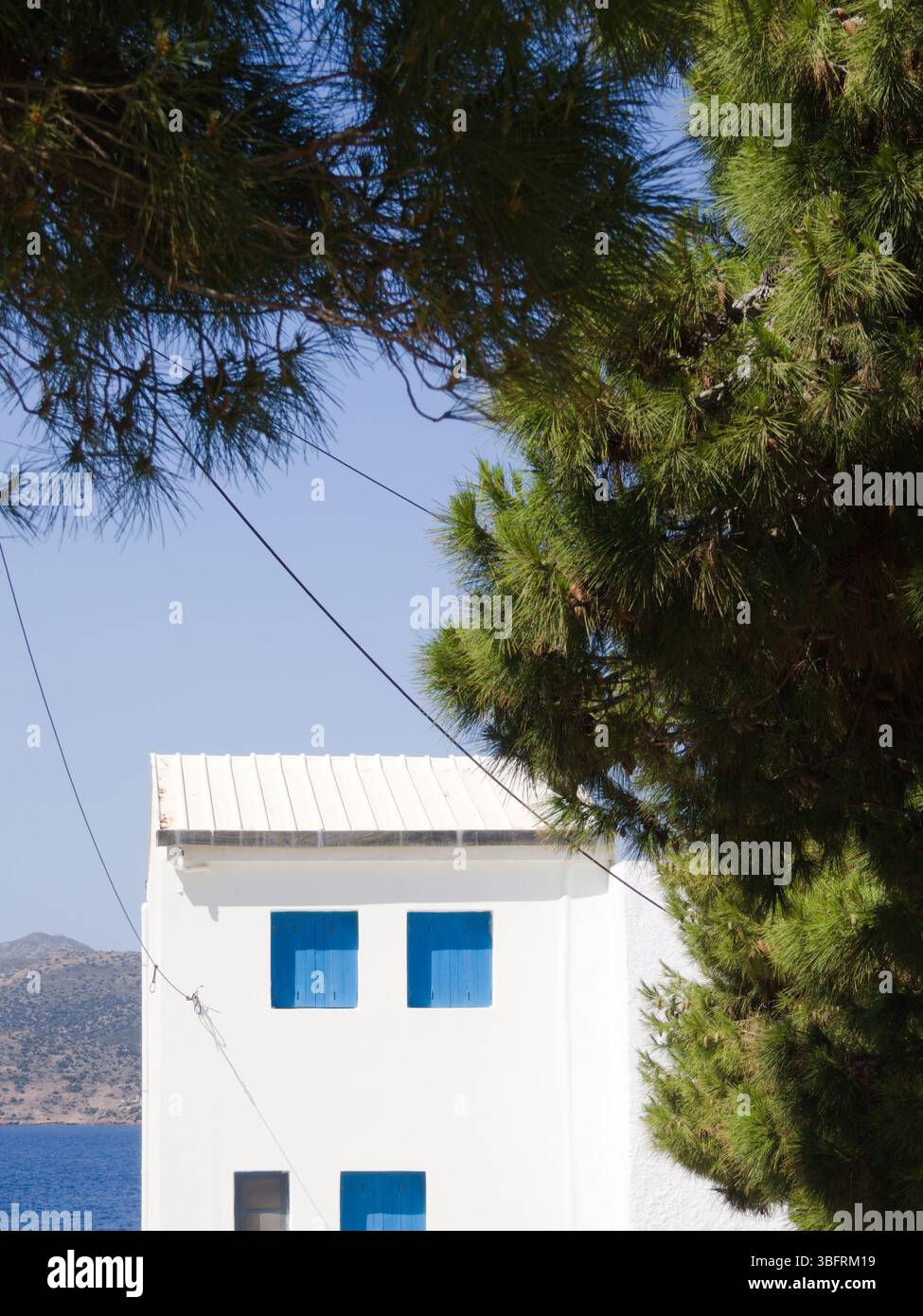 Casa dipinta di bianco con persiane azzurre, rami di pino, mare e costa sullo sfondo sotto il cielo limpido. Tranquillo scenario dell'isola greca. Foto Stock