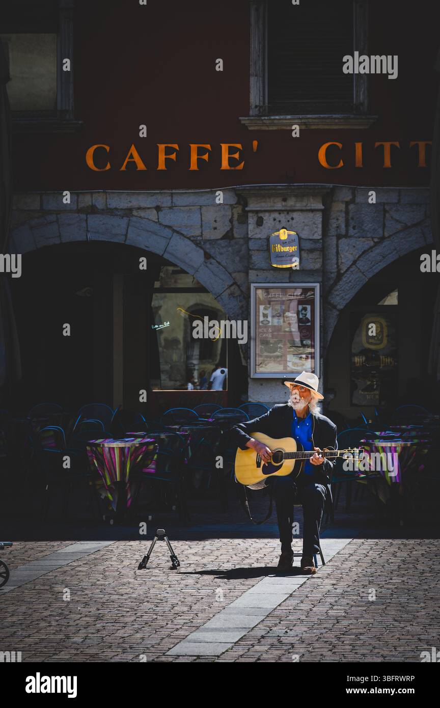 Un busker si siede al sole del mattino a Riva del Garda in Italia cantando e suonando la sua chitarra. Foto Stock
