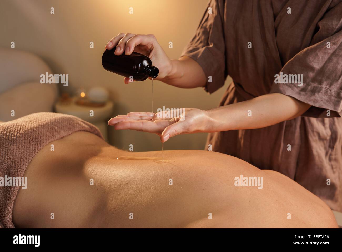 Il terapista versa olio per massaggi prima del trattamento corpo in un'atmosfera rilassante con toni caldi e luci soffuse. Foto Stock