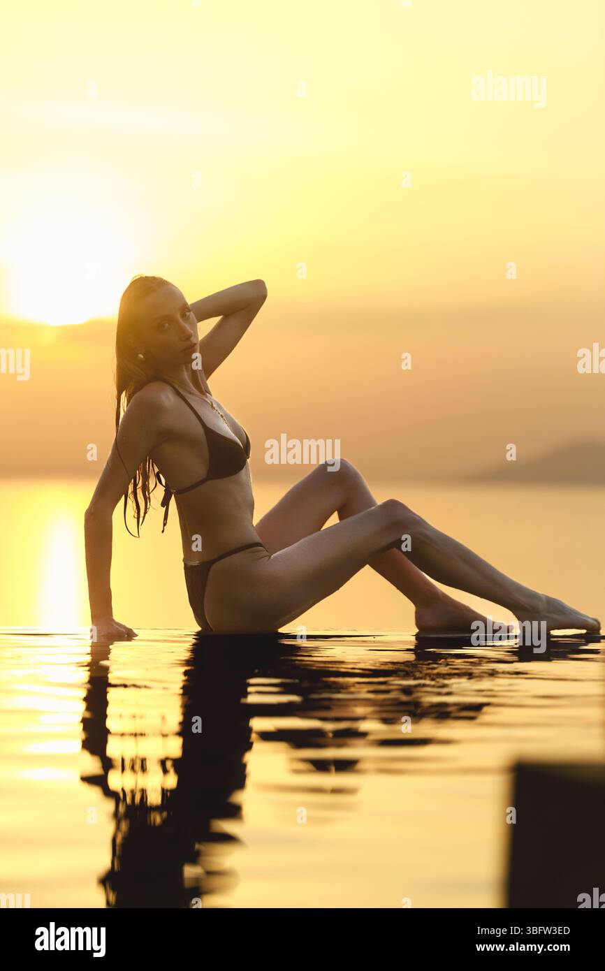 Una donna bionda degli anni '20 si mette in posa sexy mentre si siede accanto a una piscina a sfioro durante l'ora del tramonto dorato. Foto Stock