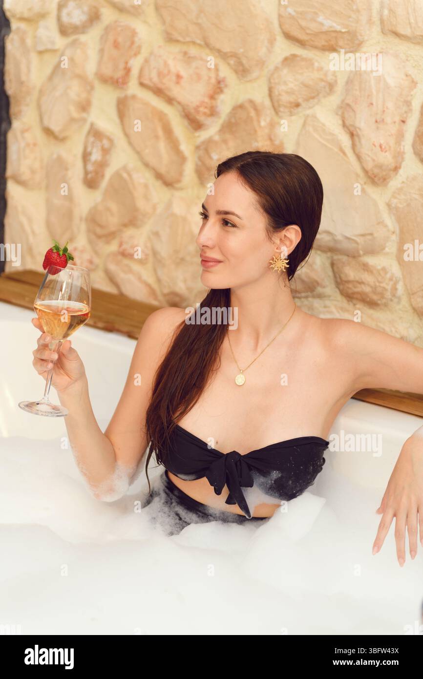 Una donna bruna degli anni '20 si rilassa in una vasca da bagno riempita di schiuma sul suo balcone dell'hotel, assaporando vino bianco tra le bollicine. Foto Stock