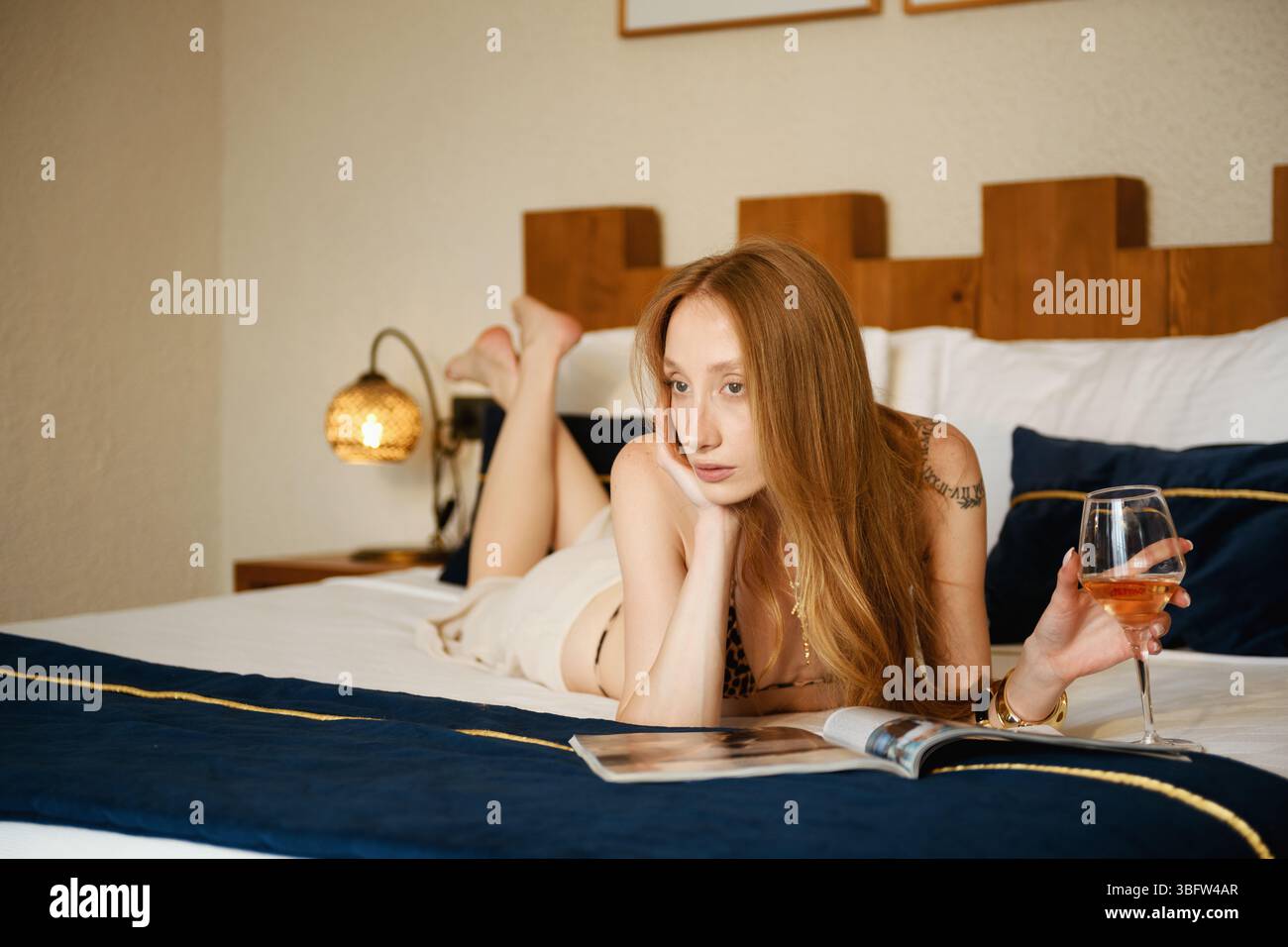 Una donna bionda degli anni '20 si rilassa sul letto del suo hotel in un bikini leopardato, posato con vino bianco e una rivista. Foto Stock