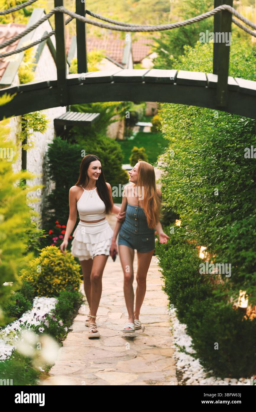 Una bruna e una donna bionda sui vent'anni sorridono mentre camminano lungo un vivace percorso verde tra le lussuose camere del resort. Foto Stock
