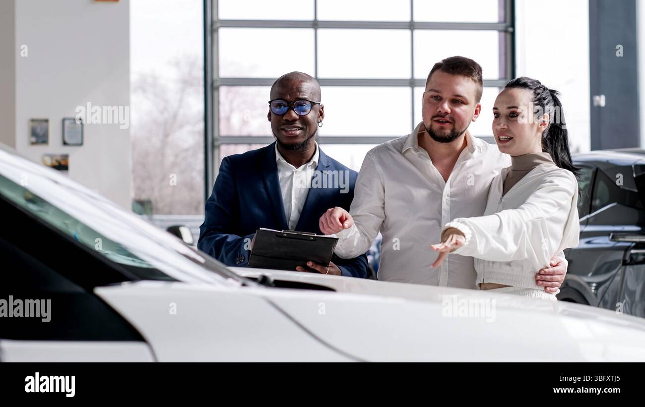 Felice coppia di sposi durante l'acquisto di una nuova e moderna auto accompagnata da un concessionario di auto nero presso una concessionaria. Foto Stock
