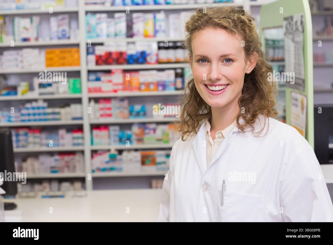 Farmacista sorridente che lavora dietro il bancone, ripiani riforniti, penna in tasca, spazio per le copie Foto Stock