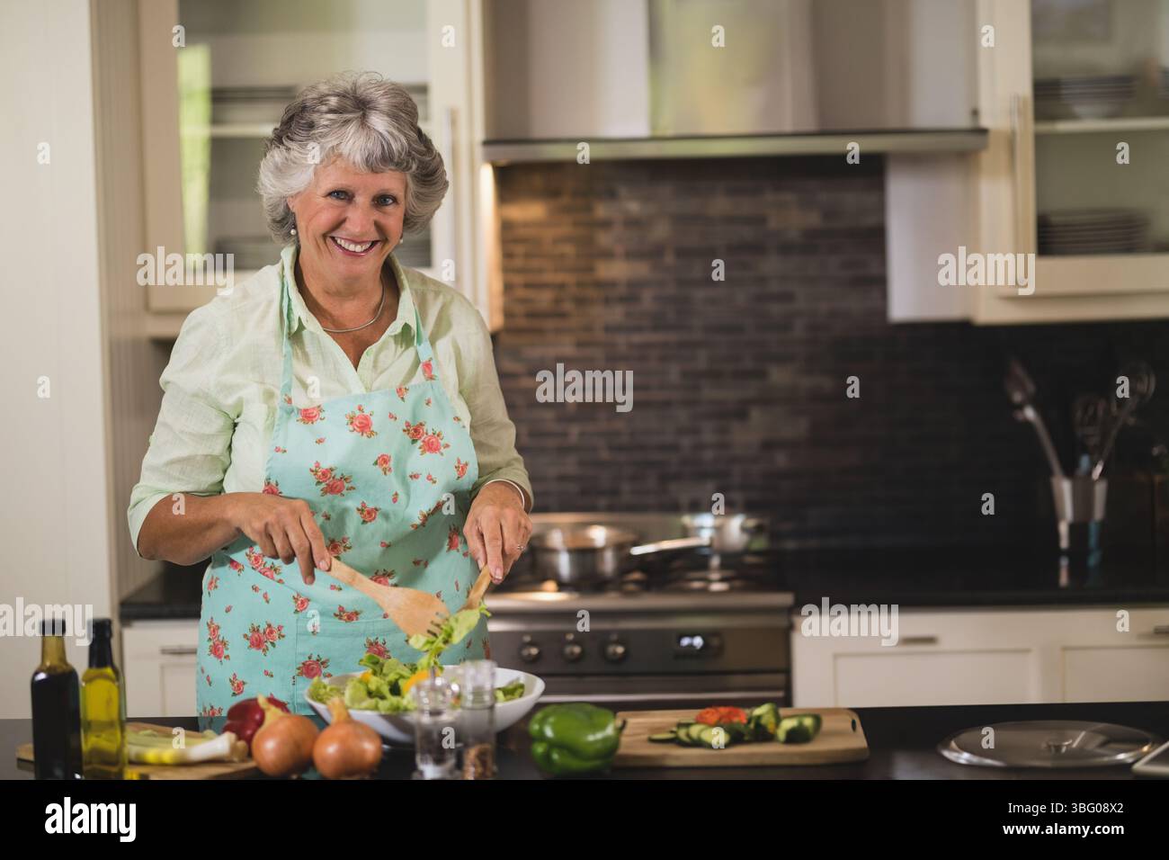 Insalata da donna anziana in ciotola bianca con cucchiaio di legno all'isola della cucina con cetrioli a fette Foto Stock