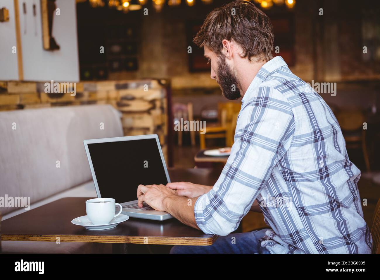 Computer portatile argentato seduto su un tavolo di legno con una tazza di caffè bianca che riposa accanto nell'accogliente caffetteria Foto Stock
