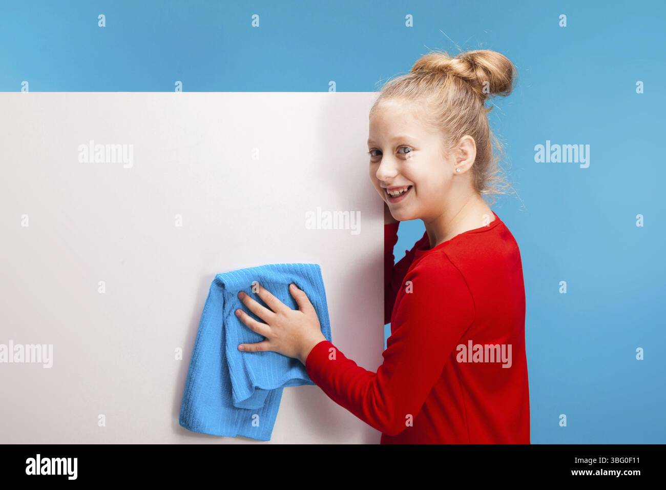 Isolata su una ragazza blu sorridente in pullover rosso con cappa dai capelli chiari, lucida il backgroung bianco con un panno blu. copyspace Foto Stock