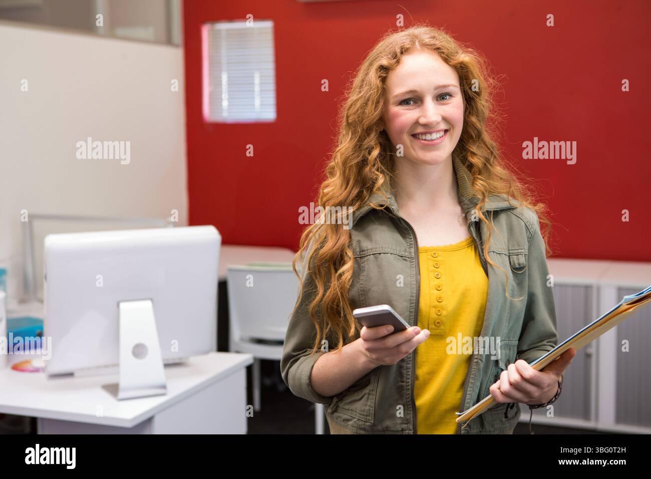 Una stagista che indossa una giacca verde, tiene in mano smartphone e appunti in un laboratorio informatico con monitor Foto Stock