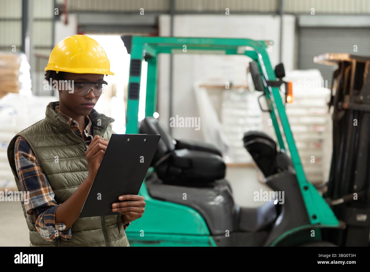 Donna che indossa gli attrezzi di sicurezza che ispeziona l'inventario del magazzino utilizzando gli appunti vicino al carrello elevatore, spazio di copia Foto Stock