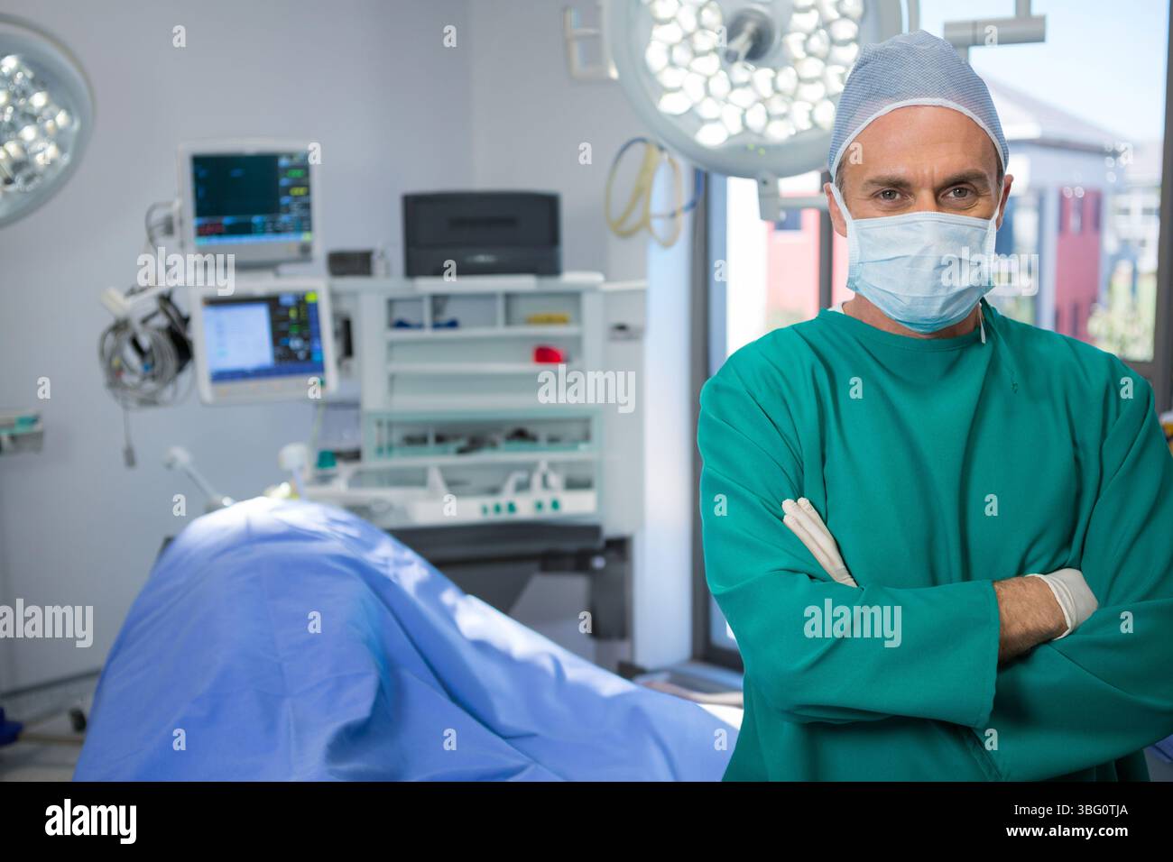 Chirurgo maschile che indossa scrub e braccia incrociate in sala operatoria sotto le luci chirurgiche, spazio di copia Foto Stock