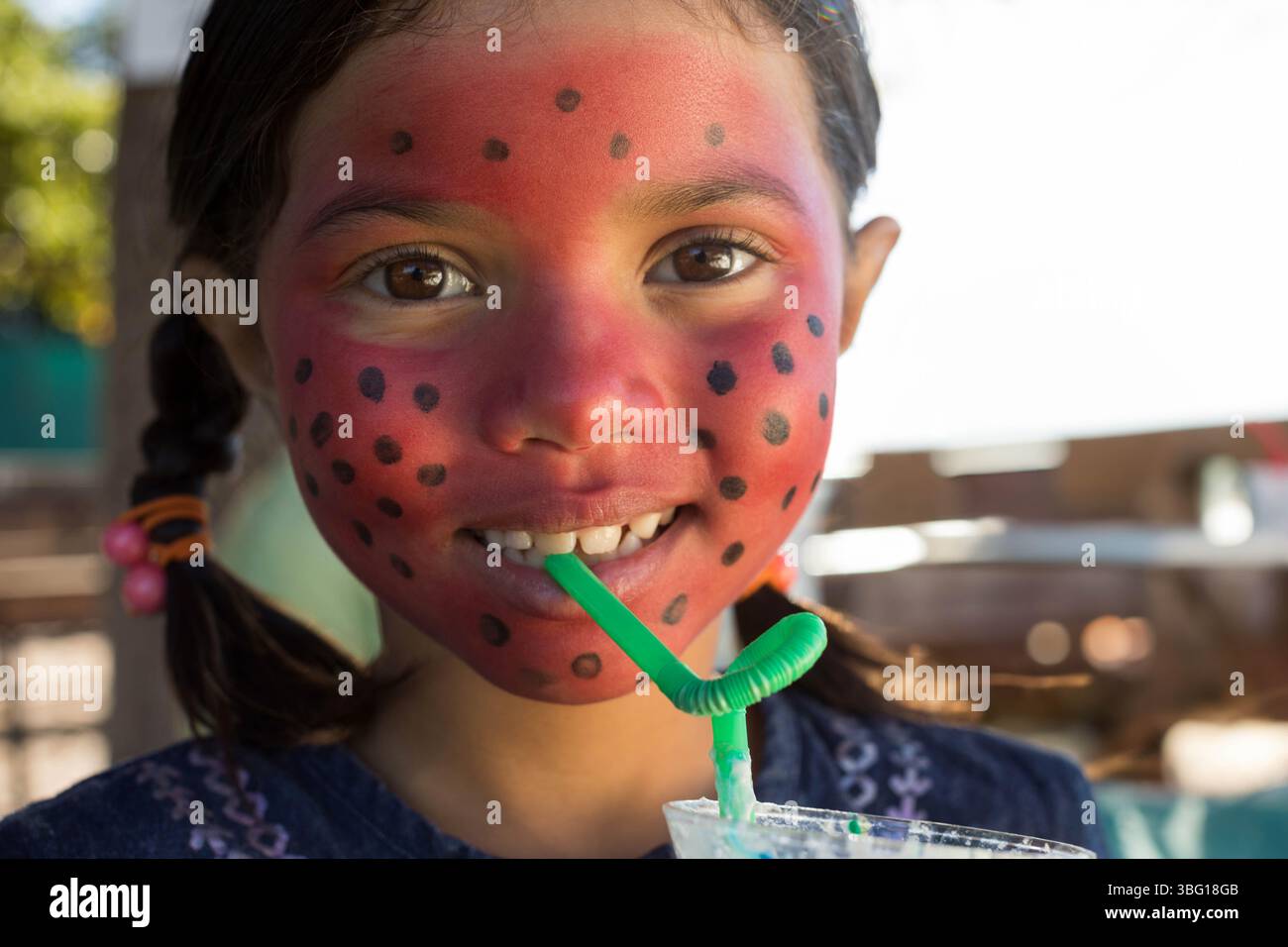 Ragazza con il viso rosso-nero macchiato in camicia sorseggiando una bevanda frizzante attraverso la paglia al tavolo di legno Foto Stock