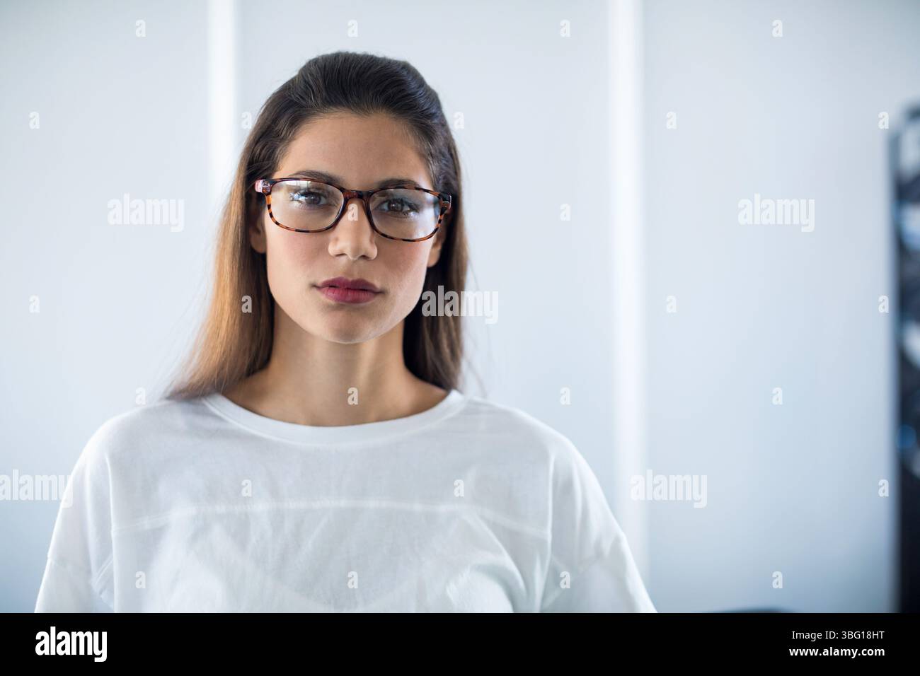 Donna in piedi contro una parete biancastra nella stanza che indossa occhiali da vista con guscio di tartaruga e camicia bianca Foto Stock