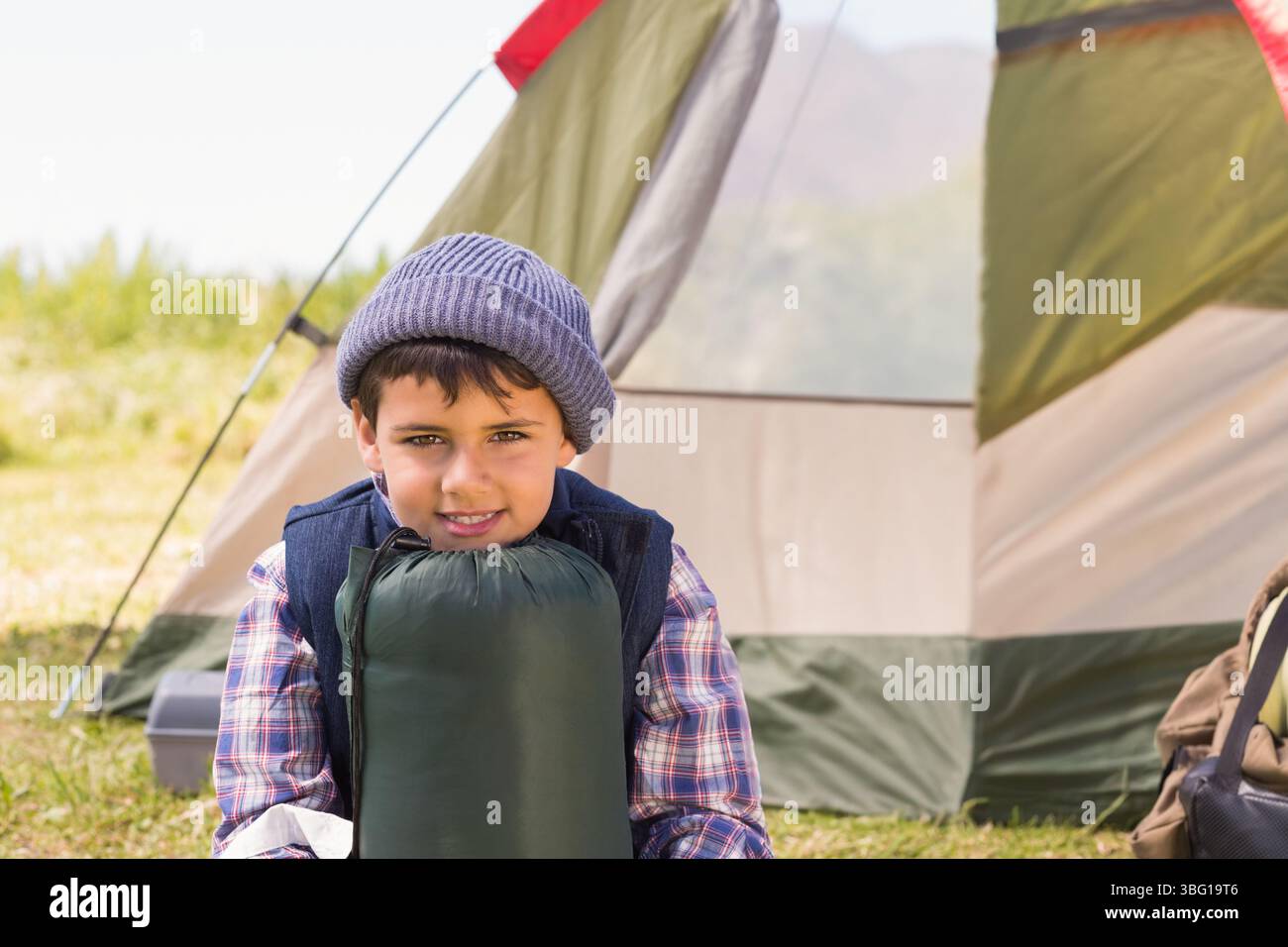 Ragazzo del Medio Oriente seduto sull'erba del campeggio tenendo in mano il sacco a pelo vicino alla tenda di tela rossa kaki Foto Stock
