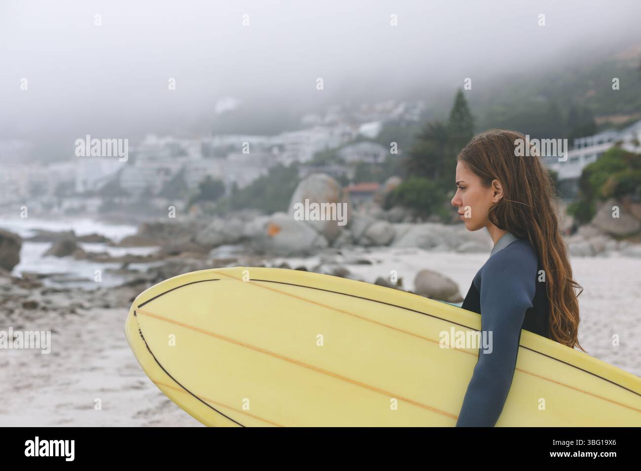 Surfista in piedi sulla spiaggia nebbiosa indossando la muta e tenendo in mano la tavola da surf gialla, spazio per copiare Foto Stock