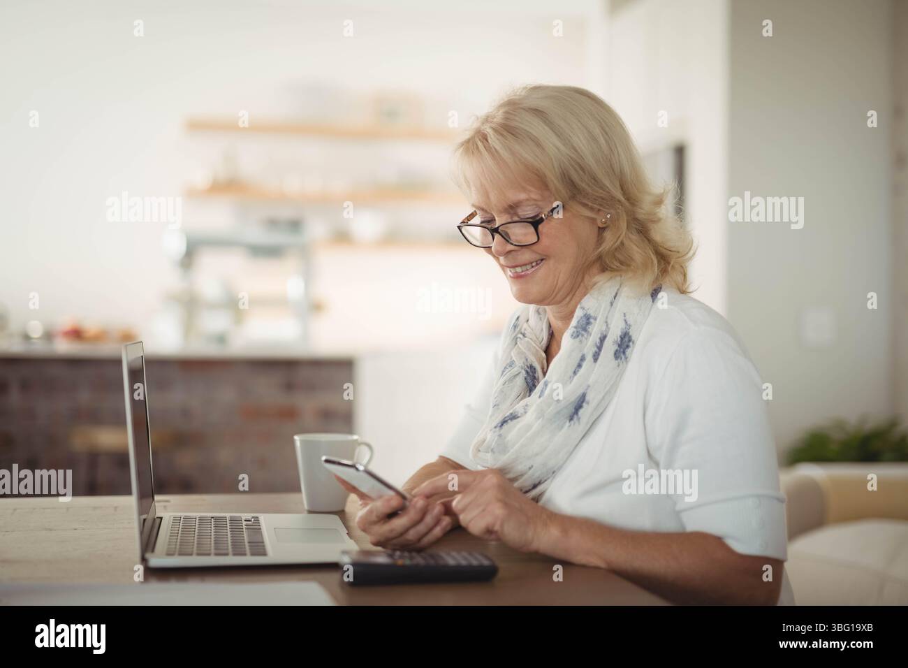 Donna anziana che sorseggia un caffè mentre utilizza un computer portatile, uno smartphone, una calcolatrice al tavolo della cucina, copia spazio Foto Stock