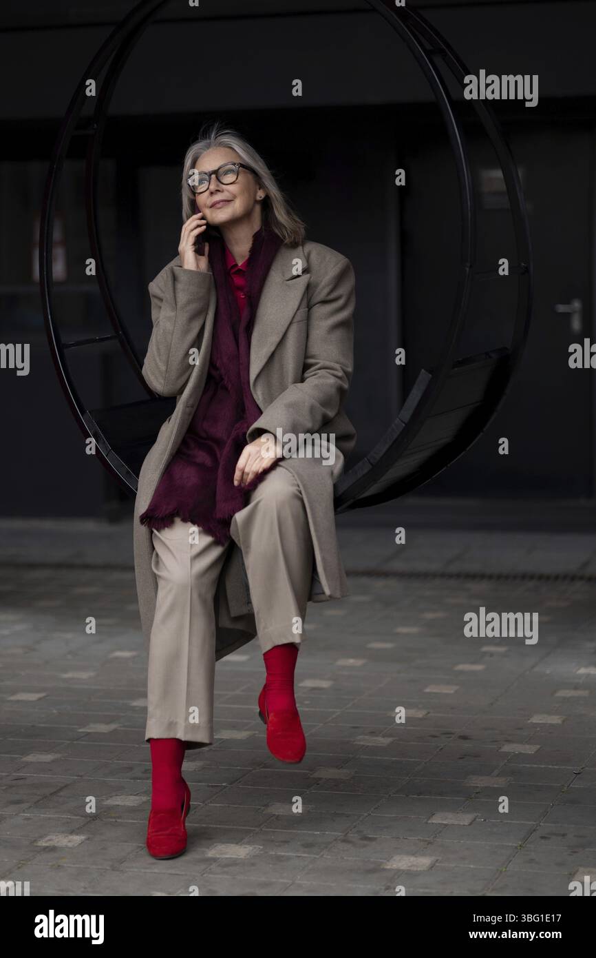 Un'anziana donna d'affari siede su un'altalena circolare fuori dal suo ufficio, impegnata in una conversazione telefonica. È vestita con un abbigliamento professionale, showcasi Foto Stock
