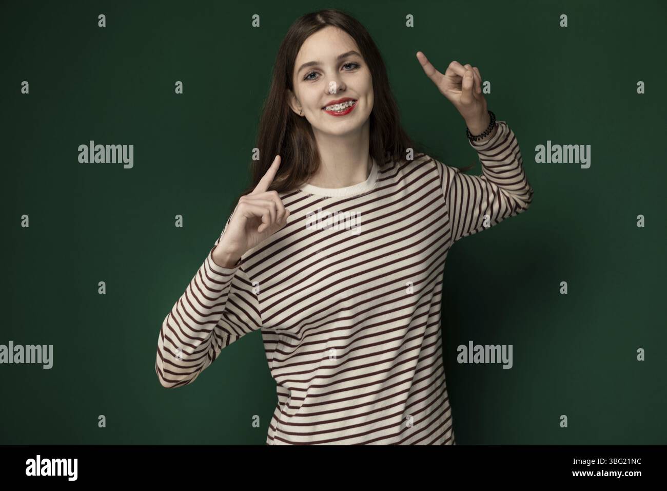 Una donna che indossa una camicia a righe posa per la fotocamera, tenendo le dita in alto con un gesto di pace. Guarda direttamente lo spettatore, con un neu Foto Stock