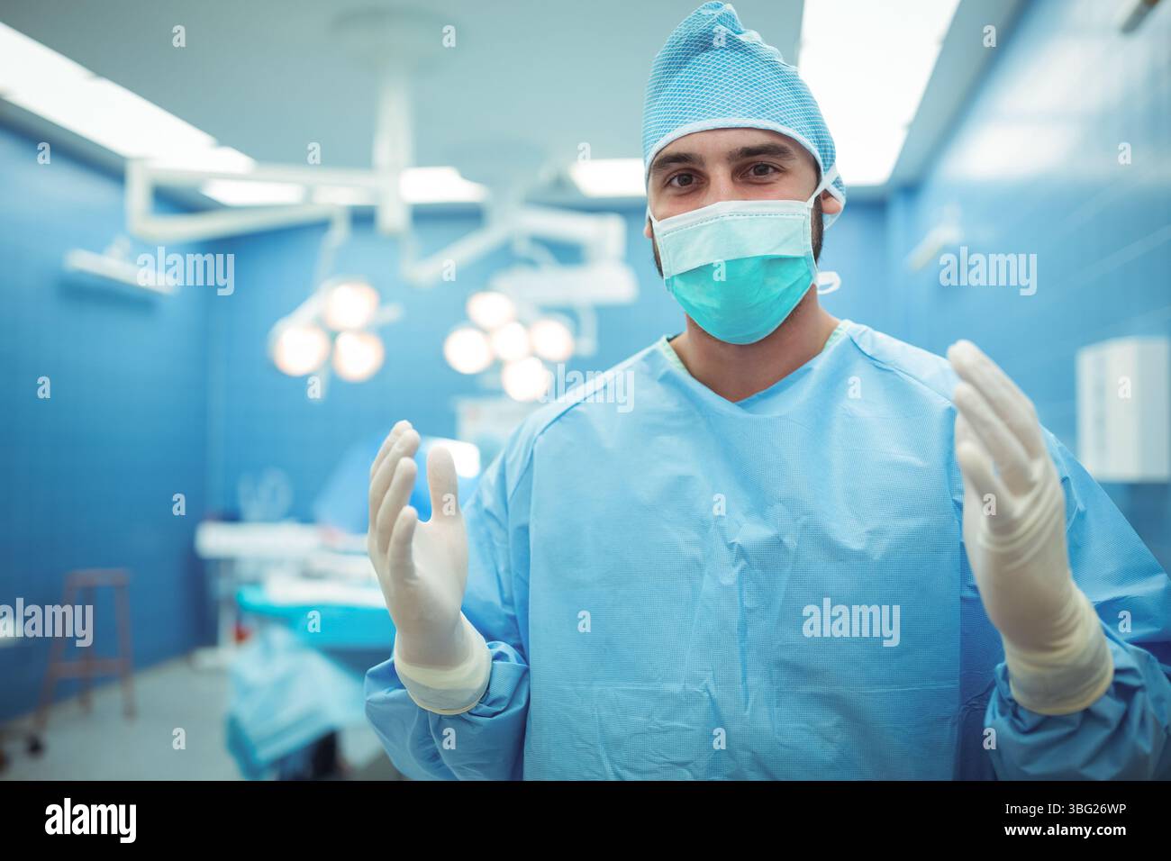 Chirurgo maschile in camice chirurgico, maschera in piedi in sala operatoria sotto le luci accanto al vassoio dello strumento Foto Stock