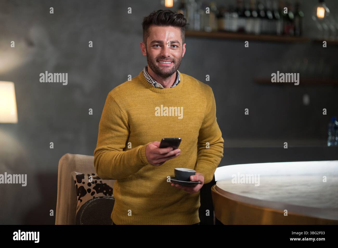 Uomo che indossa un maglione di senape che tiene in mano lo smartphone e una tazza di caffè al bancone del caffè vicino a scaffali di legno Foto Stock