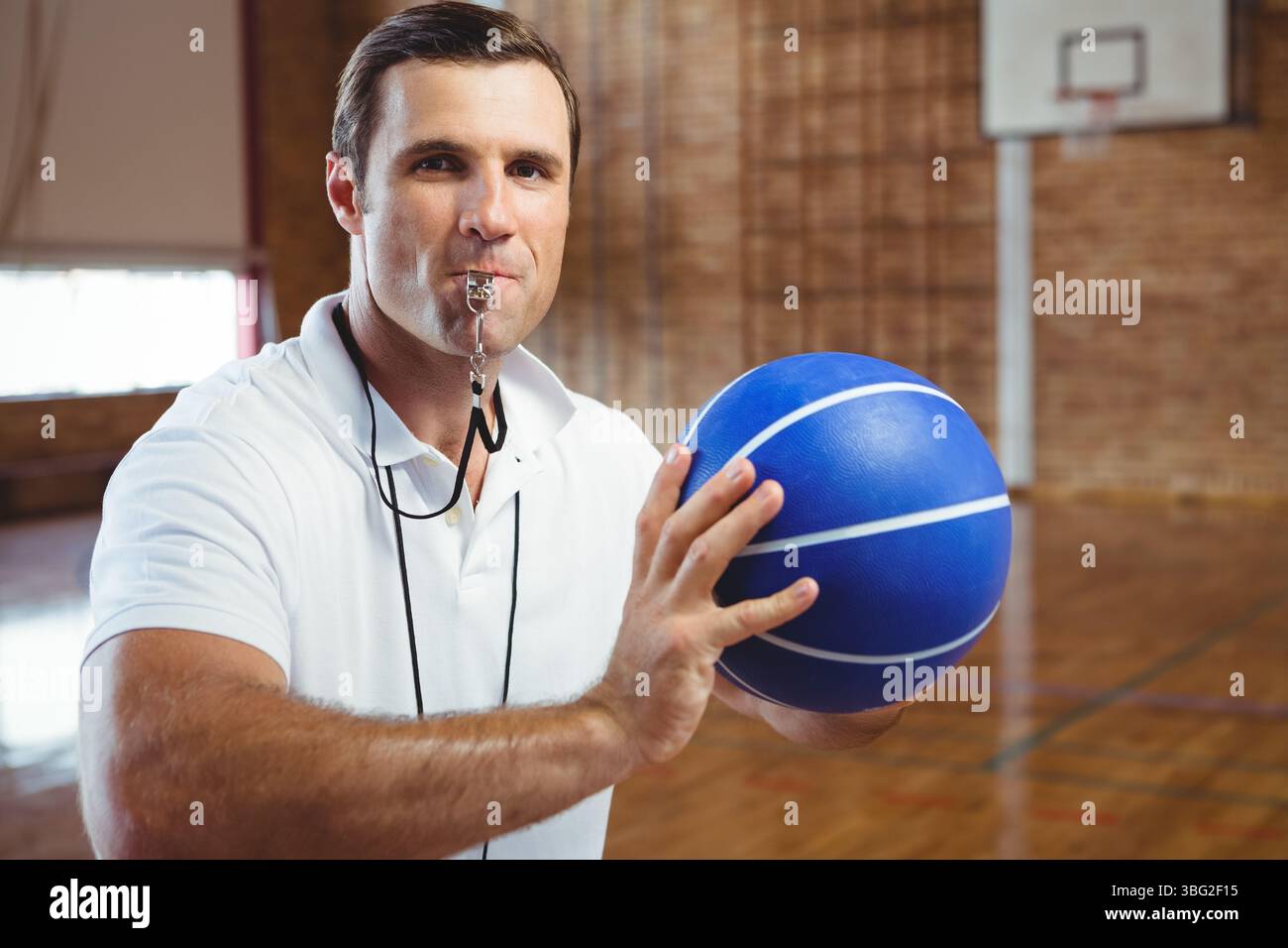 Arbitro maschile adulto maturo con pallacanestro blu e fischio in piedi sul campo da ginnastica Foto Stock