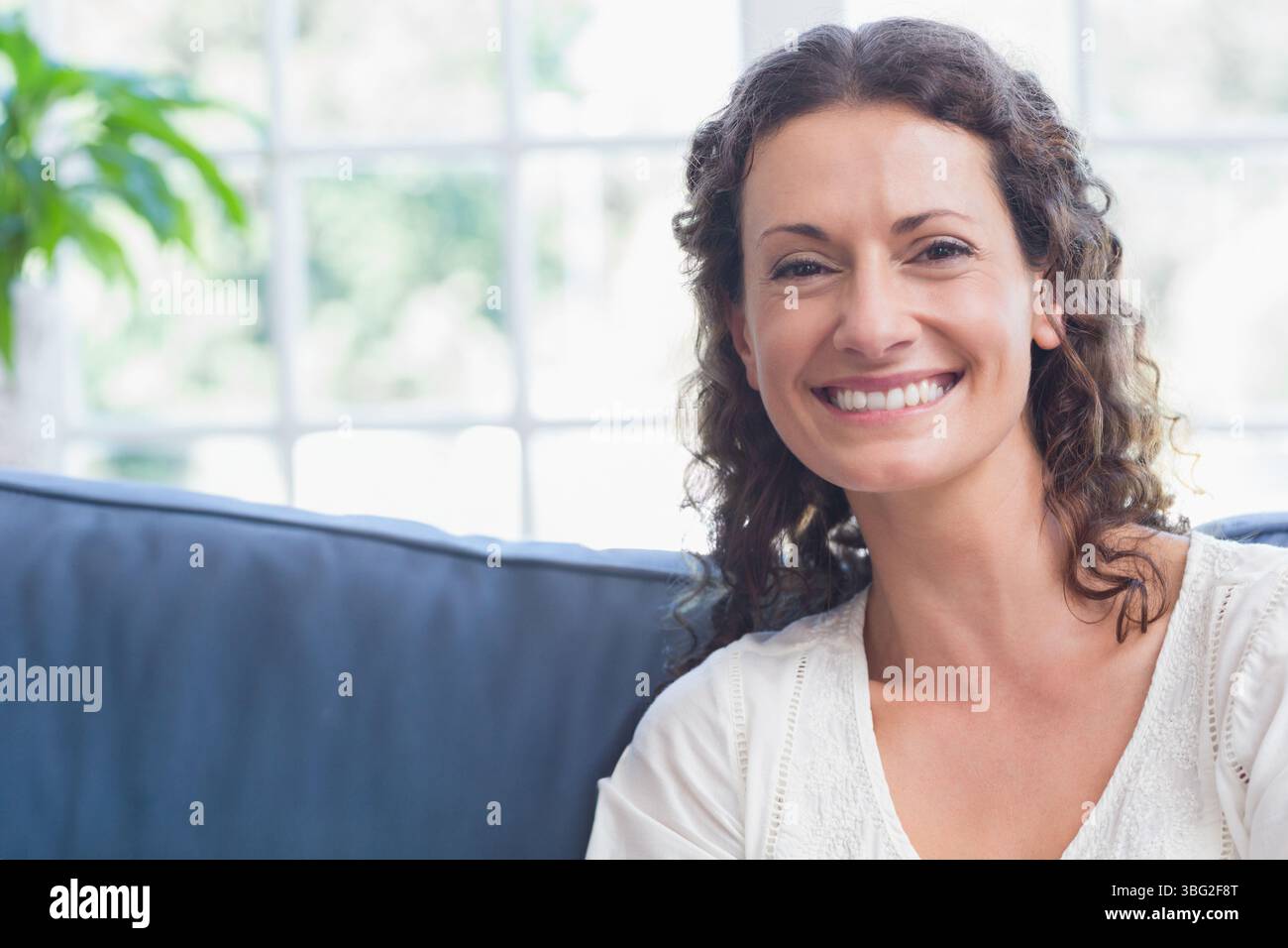 Donna seduta su un divano blu scuro vicino a una finestra a più pannelli nel soggiorno, pianta in vaso, spazio per copiare Foto Stock