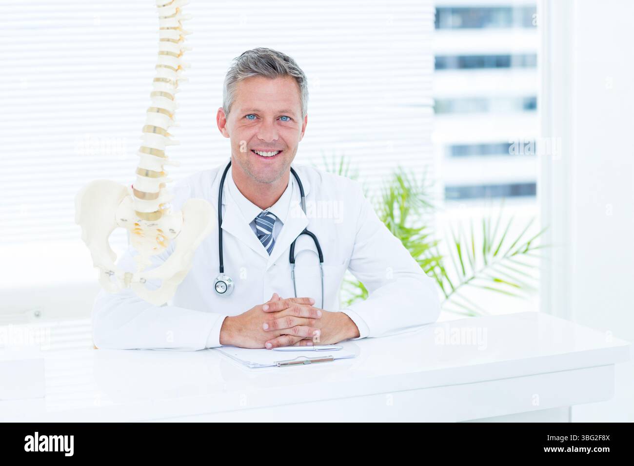 Medico maschio maturo che indossa un cappotto bianco e uno stetoscopio seduto alla scrivania dell'ufficio esaminando il modello della colonna vertebrale Foto Stock