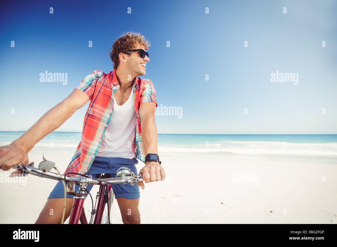 Un uomo adulto che va in bicicletta lungo la costa sabbiosa con onde oceaniche, con occhiali da sole e smartwatch Foto Stock