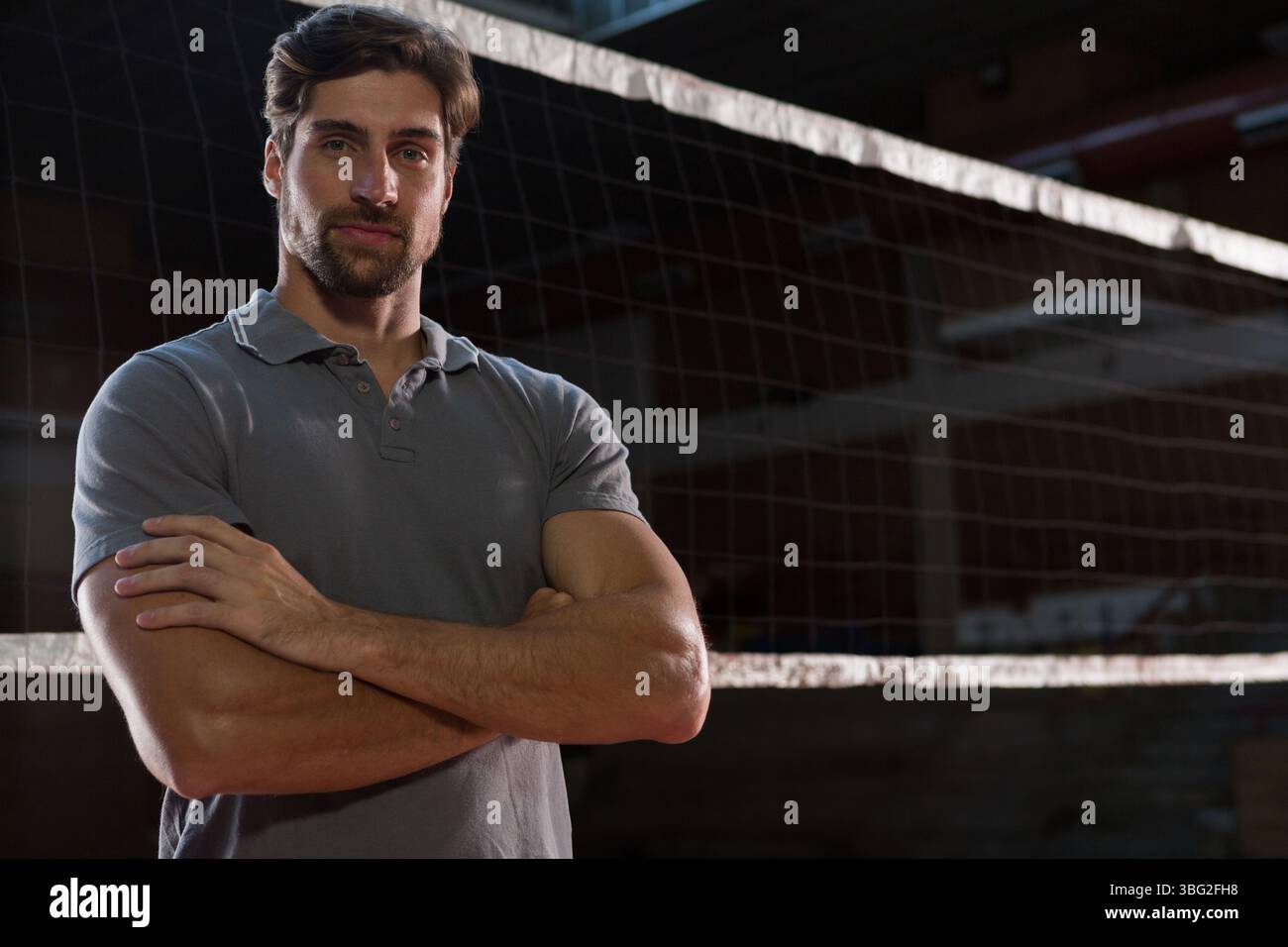 Uomo in piedi con le braccia incrociate e indossa una polo grigia sui pali della rete da pallavolo nella sala dello sport Foto Stock